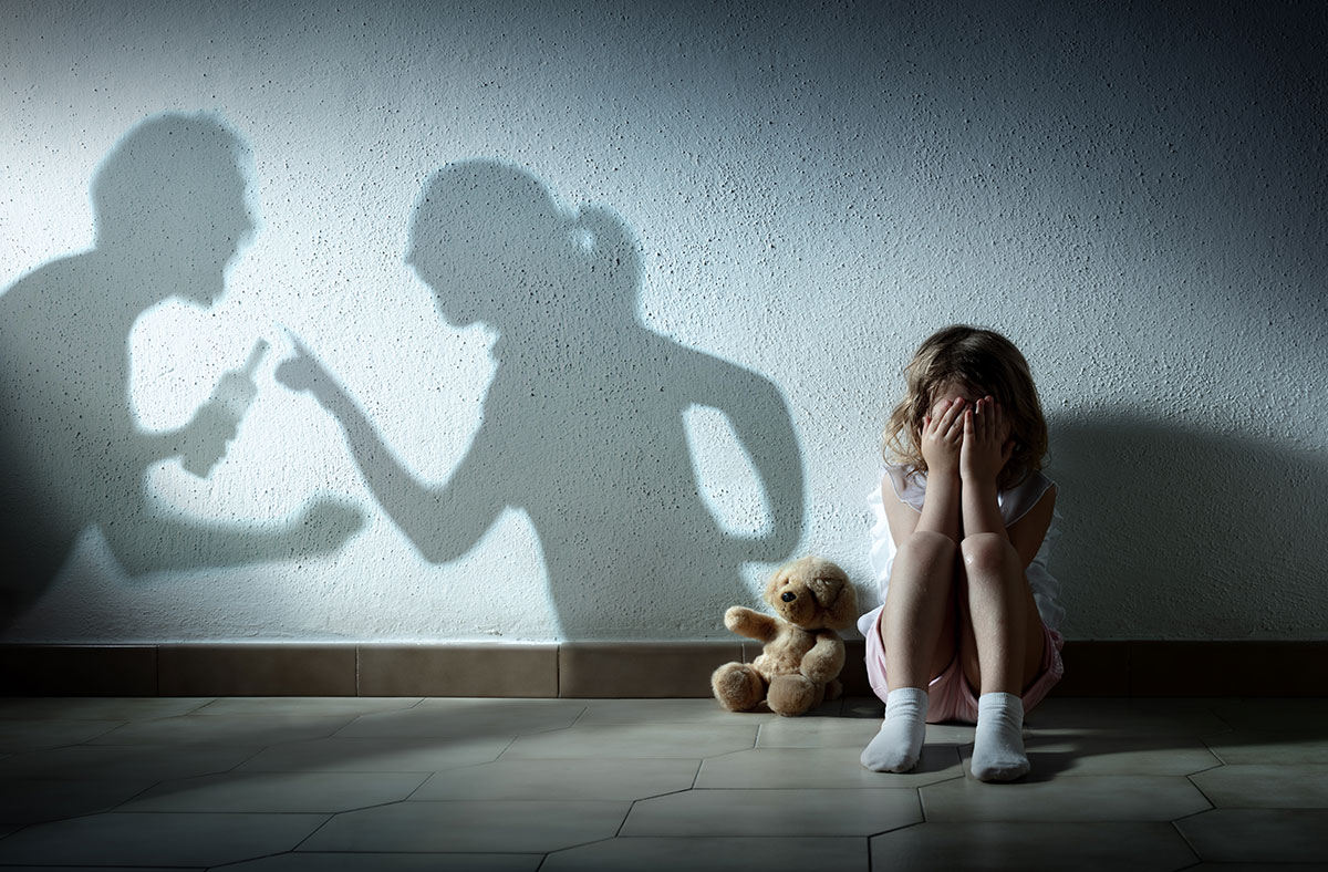 Ein Kind sitzt weinend, mit verschränkten Händen vor dem Gesicht auf dem Boden. Neben dem Kind sitzt ein kleiner Teddybär. Daneben sieht man die Schatten von zwei erwachsenen Personen an der Wand.  Die beiden streiten miteinander und halten Flaschen mit Alkohol in Händen.